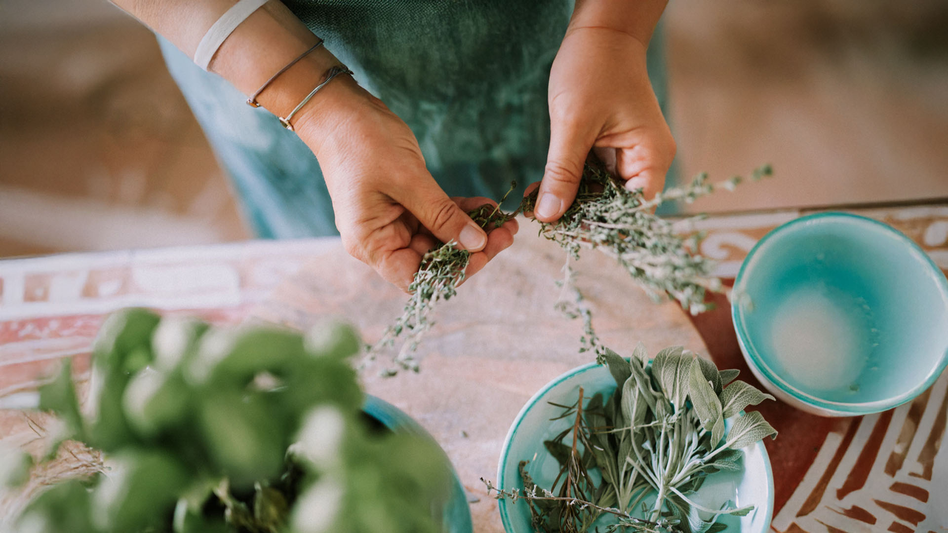 Hands holding and separating fresh herbs above bowls filled with other herbs on a patterned table.