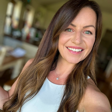 Smiling woman with long brown hair and green eyes wearing a white sleeveless top and star necklace indoors.
