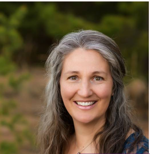 Smiling middle-aged woman with long gray and dark hair standing outdoors with blurred green foliage background.