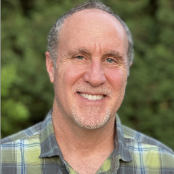 Smiling middle-aged man with short gray hair and a light beard, wearing a plaid shirt, standing outdoors with green foliage background.
